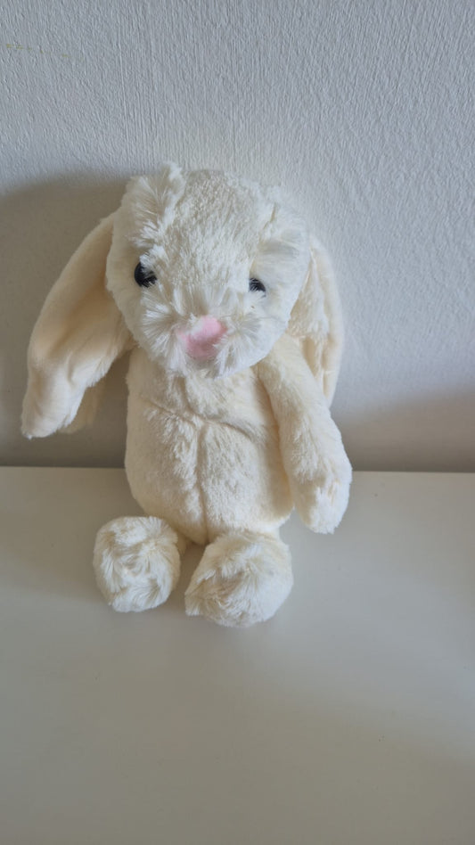 Zachte crèmekleurige pluche konijn knuffel met hangende oren, zwarte ogen en een roze neusje, zittend tegen een witte muur.